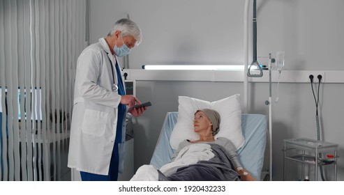 Doctor Telling Results Of Medical Tests To Cancer Patient In Hospital Ward. Senior Oncologist In Safety Mask Discussing Treatment With Sick Aged Woman Lying In Bed In Clinic