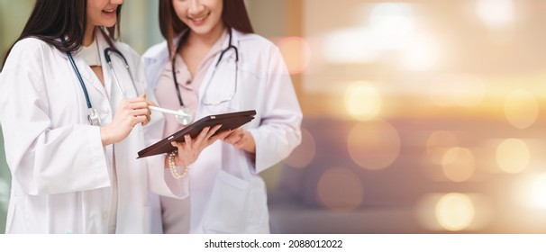 Doctor in a uniform, a stethoscope and a female assistant stand together in a hospital corridor. Hold the data file and tablet showing the patient's symptoms. Asian and European women. - Powered by Shutterstock