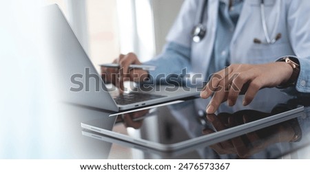 Doctor using digital tablet and working on laptop computer at doctor's office in hospital, close up. Healthcare and medicine, medical technology background
