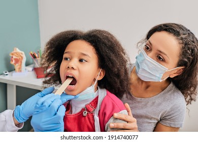 Doctor using inspection spatula to examine African American girl's throat. Little girl with her mom at pediatrician appointment