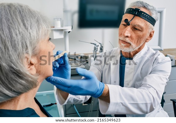 Doctor using inspection spatula\
examines senior woman throat. ENT doctor consultation, throat\
exam