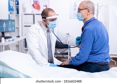 Doctor using stethoscope to listen senior man heart during examination in hospital room and wearing visor as safety precaution against coronavirus. Medical control for infections, disease.