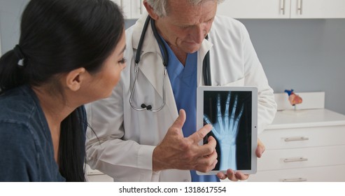 Doctor Using Tablet Computer With X-ray Of Hand To Talk About Recovery With Female Patient. Senior Caucasian Male Medical Professional With Screen Demonstrating Arthritis To Woman