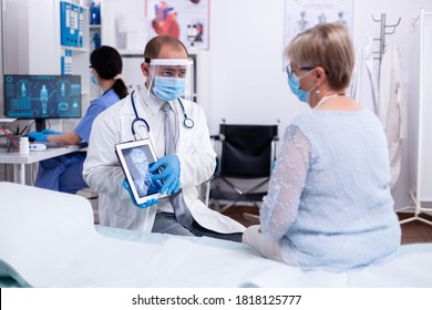 Doctor using tablet pc during examination of x-ray for older patient in hospital room and wearing face mask against coronavirus pandemic. Medical examination for infections, disease and diagnosis.