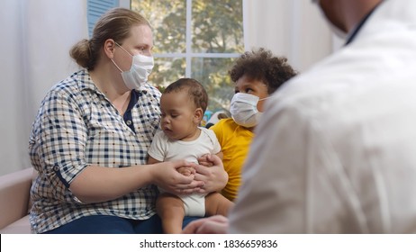 Doctor Visiting Multiethnic Family With Little Children Wearing Protective Mask. Pediatrics Consulting Foster Mother Holding Baby Son With Toddler Boy Sitting Behind On Couch At Home