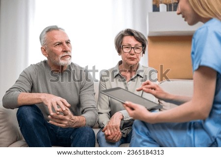 Doctor visits elderly patients at home, communicates with a senior family couple, making online notes on a tablet, using app for adding diagnosis information to the medical card