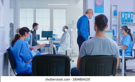 Doctor With Visor Against Coronavirus And Patient In Examination Room. Senior Man With Face Mask Talking With Nurse At Reception. Assistant And Disabled Woman With Walking Frame In Waiting Area.