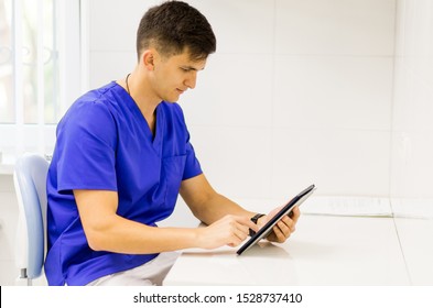 Doctor wearing blue medical uniform working with a tablet in a dentist office. Stomatologist filling in CRM form on tablet. Technologies in healthcare.
