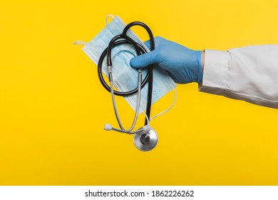 A doctor wearing a blue protective glove holds a stethoscope and a surgical mask against COVID-19 SARS-CoV-2 infections. Isolated on a yellow background.