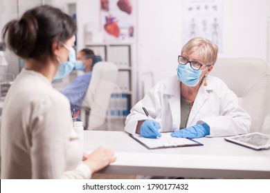 Doctor Wearing Protection Mask Against Covid Taking Notes During Consultation With Patient In Medical Clinic. Nurse Wearing Blue Uniform While Working On Computer.