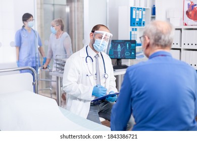 Doctor wearing visor as safety precaution against coronavirus outbreak during consultation of old man in cabinet. Modern private clinic or hospital. Practitioner physician appointment - Shutterstock ID 1843386721