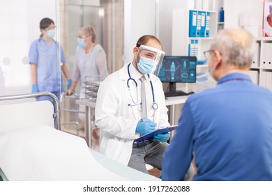 Doctor wearing visor as safety precaution in the course of consultation of senior man. Physician practitioner in the course of private modern clinic appointment during coronavirus pandemic. - Shutterstock ID 1917262088