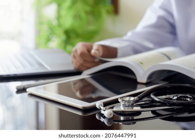 Doctor In White Gown Coat Reading Book With Digital Tablet, Laptop Computer And Stethoscope On The Desk In Medical Room At Clinic Or Hospital. Online Medical Education Concept.