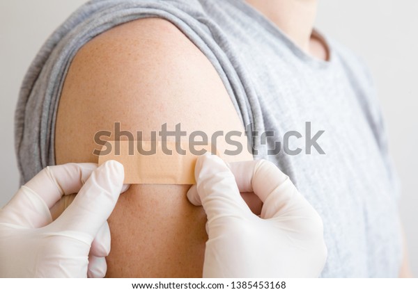 Doctor\
in white rubber protective gloves putting adhesive bandage on young\
man\'s arm after scratch on skin or injection of vaccine. First aid.\
Medical, pharmacy and healthcare concept.\
Closeup.