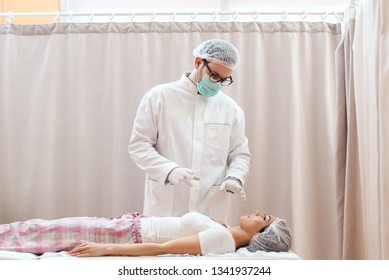 Doctor In White Uniform, With Mask On Face And Medical Cap On Head Calming Female Patient Before Surgery. Patient Lying Down On Hospital Bed.