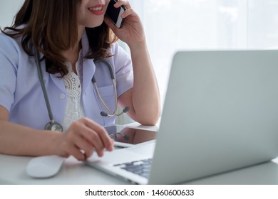 Doctor Woman, With White Gown And Stethoscope, Sitting In White Table Smiling, Talking On The Phone And Typing In Portable Laptop Computer, In Desktop Office