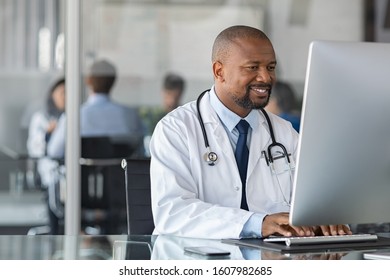 Doctor at work in office using computer with copy space in modern clinic. African happy specialist wearing white coat and sitting at desk while medical team having meeting in background.