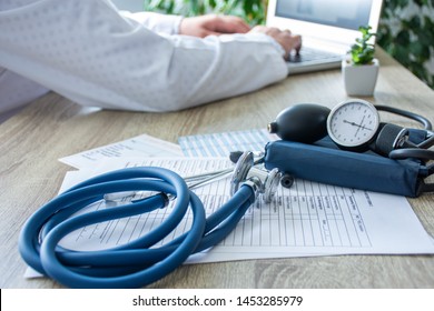 Doctor at work on the computer in the blurry background, in the foreground - a stethoscope with sphygmomanometer on medical documents on the table. Scene of doctor working in a modern medical office