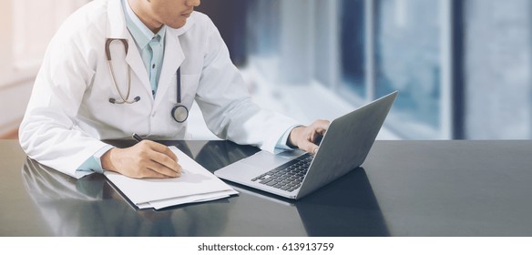 Doctor Working With Laptop Computer And Writing On Paperwork. Hospital Background.