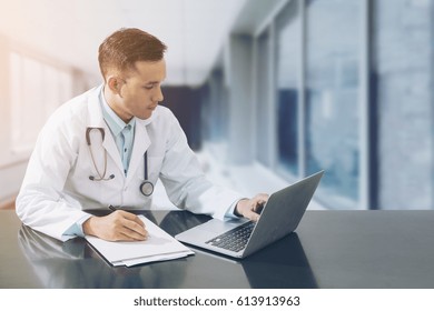 Doctor Working With Laptop Computer And Writing On Paperwork. Hospital Background.