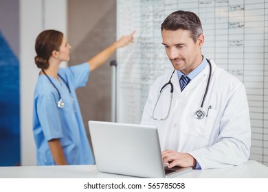 Doctor Working On Laptop With Colleague Pointing At Chart In Hospital