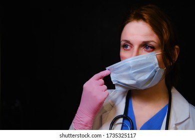 A Doctor With Wounds On His Face Takes Off A Medical Mask. Nurse On A Black Background With Mask Marks, Closeup. Concept Of Overworking Doctors During The Crisis With Coronavirus