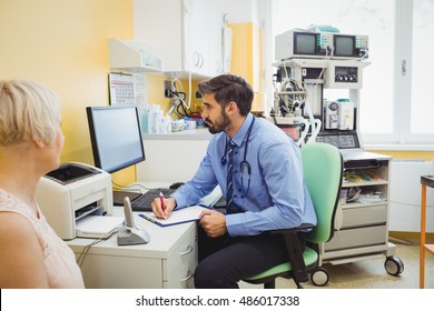 Doctor Writing On Clipboard While Looking At Computer In The Hospital