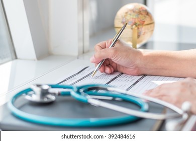 Doctor Writing On Medical Health Care Record, Patients Discharge, Or Prescription Form Paperwork In Hospital Clinic With Physician's Stethoscope On Office Desk