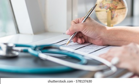 Doctor Writing On Medical Health Care Record, Patients Discharge, Or Prescription Form Paperwork In Hospital Clinic Office With Physician's Stethoscope On Desk