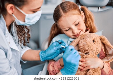 At the doctor's appointment. A child is sitting in a dental chair and holding a toy rabbit and a nurse is playfully examining the toy. Approach to the patient. - Powered by Shutterstock