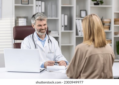 Doctor's appointment. A senior man doctor sits in the office at a table with a laptop and receives patients, consults and talks with a young woman. - Powered by Shutterstock