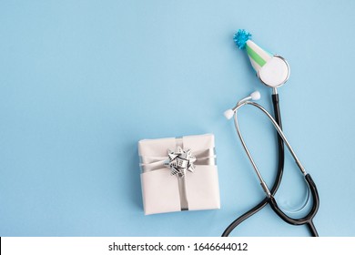 Tag des Arztes und Weltgesundheitstag. Medizinisches Stethoskop in festlicher Kappe und Geschenk auf hellblauem Hintergrund. Kreative medizinische Postkarte. Draufsicht, flache Lage, Kopienraum