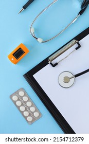 Doctors Desk, Table. Medical Accessories. Workplace Desktop Cardiologist, Therapist. Healthcare Concept. Stethoscope, Pulse Oximeter, Pills, Medicines, Paper Blank Sheet On Blue Background. Vertical