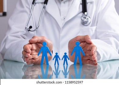 Doctor's Hand Protecting Blue Family Figure Cutout On Reflective Desk