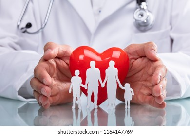 Doctor's Hand Protecting Red Heart With Family Figure On Reflective Desk