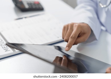 A doctor's hand is writing on a medical record form on a clipboard, with a stethoscope and tablet in the background. - Powered by Shutterstock