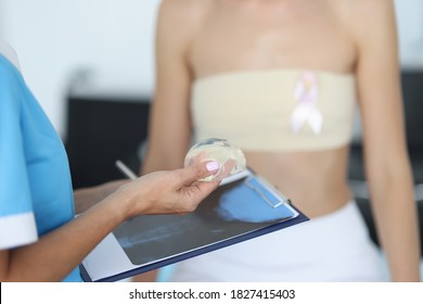 Doctor's Hands Are Holding Breast Implant In Front Of Clipboard With Patient In Background. Mammaplasty Breast Correction Concept.