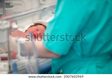 The doctor's hands in rubber gloves are holding the head of a newborn baby who lies in the medical box.