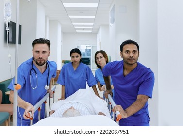 Doctors And Nurses Wheeling Elderly Patient In Gurney On Hospital Corridor At Emergency Department. Work Of Emergency Medical Team And Paramedics