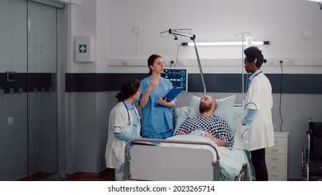 Doctors Team Checking Sick Man Discussing Sickness Symptom Monitoring Medical Recovery In Hospital Ward. Patient Resting In Bed Waiting Disease Treatment During Rehabilitation Appointment