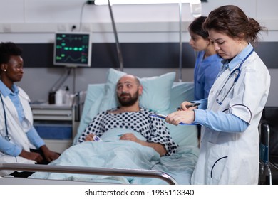 Doctors Team Monitoring Man Examining Illness Symptom Monitoring Medical Recovery In Hospital Ward. Sick Patient Resting In Bed Waiting Sickness Treatment During Rehabilitation Appointment