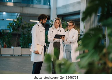 Doctors using digital tablet, collaborating on patient information in bright, modern hospital lobby with plants, showcasing teamwork and technology in healthcare