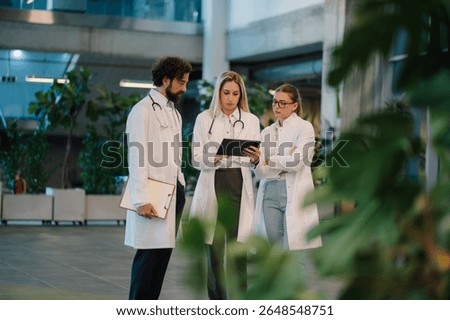 Doctors using digital tablet, collaborating on patient information in bright, modern hospital lobby with plants, showcasing teamwork and technology in healthcare