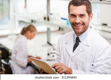 Documenting the result of experiments. Smiling young male scientist holding digital tablet and looking at camera while his female colleague working in the background