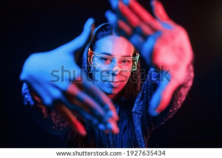 Does gestures by hands. Portrait of young girl that is in eyeglasses in blue neon lighting.