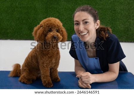Dog groomer smiling while posing with a freshly groomed toy poodle on a table.