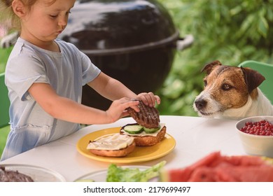 Dog Looking How Little Girl Making Burger At BBQ Party On Sunny Summer Day