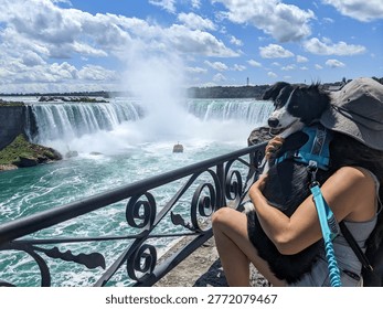 A dog looks at the camera while being held by a woman overlooking Niagara Falls with a maid of the mist boat on the river during a sunny summer day.