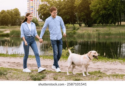 Dog-Friendly Date Concept. Two young people walking dog on a leash in park near lake or river, holding hands