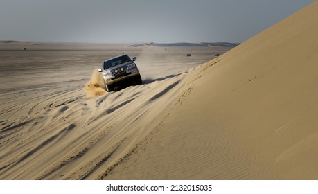 Doha, Qatar: Circa 2021: A Speeding Sports Utility Vehicle Driving Along A Sloped Dune Face Splashing Sand From Wheels. Adventurous People Love Speed And Sand Dune Bashing.
